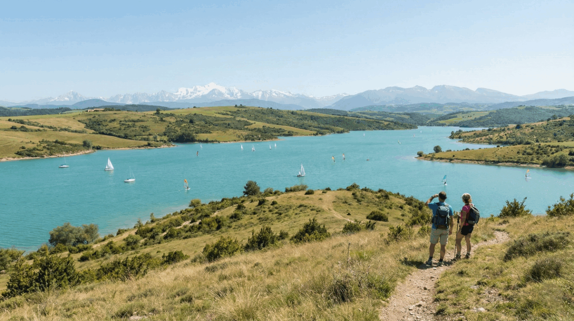 Vue panoramique sur le Lac de la Ganguise aux eaux turquoise avec activités nautiques et la chaîne des Pyrénées en arrière-plan.
